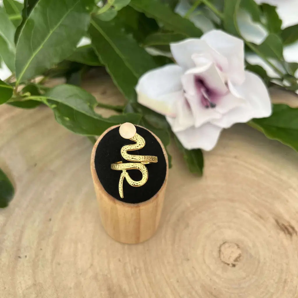 Bague ajustable Serpent en métal doré présentée sur un support en bois avec fond noir et fleurs blanches