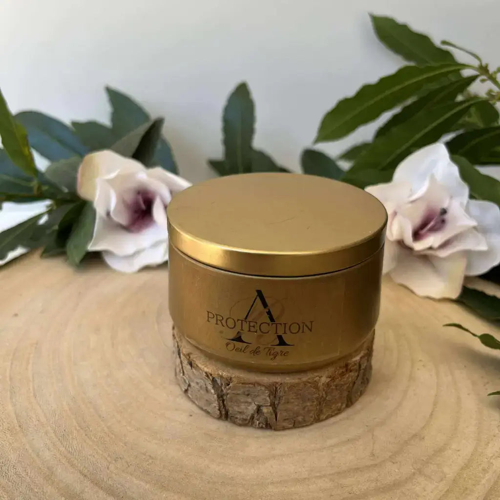 Bougie Protection en Œil de Tigre Aura de Roche en boîte dorée posée sur rondin de bois avec fleurs et feuillage