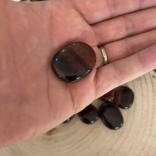 Galet Œil de Taureau poli brun rouge posé dans une main, autres galets brillants sur planche en bois Aura de Roche