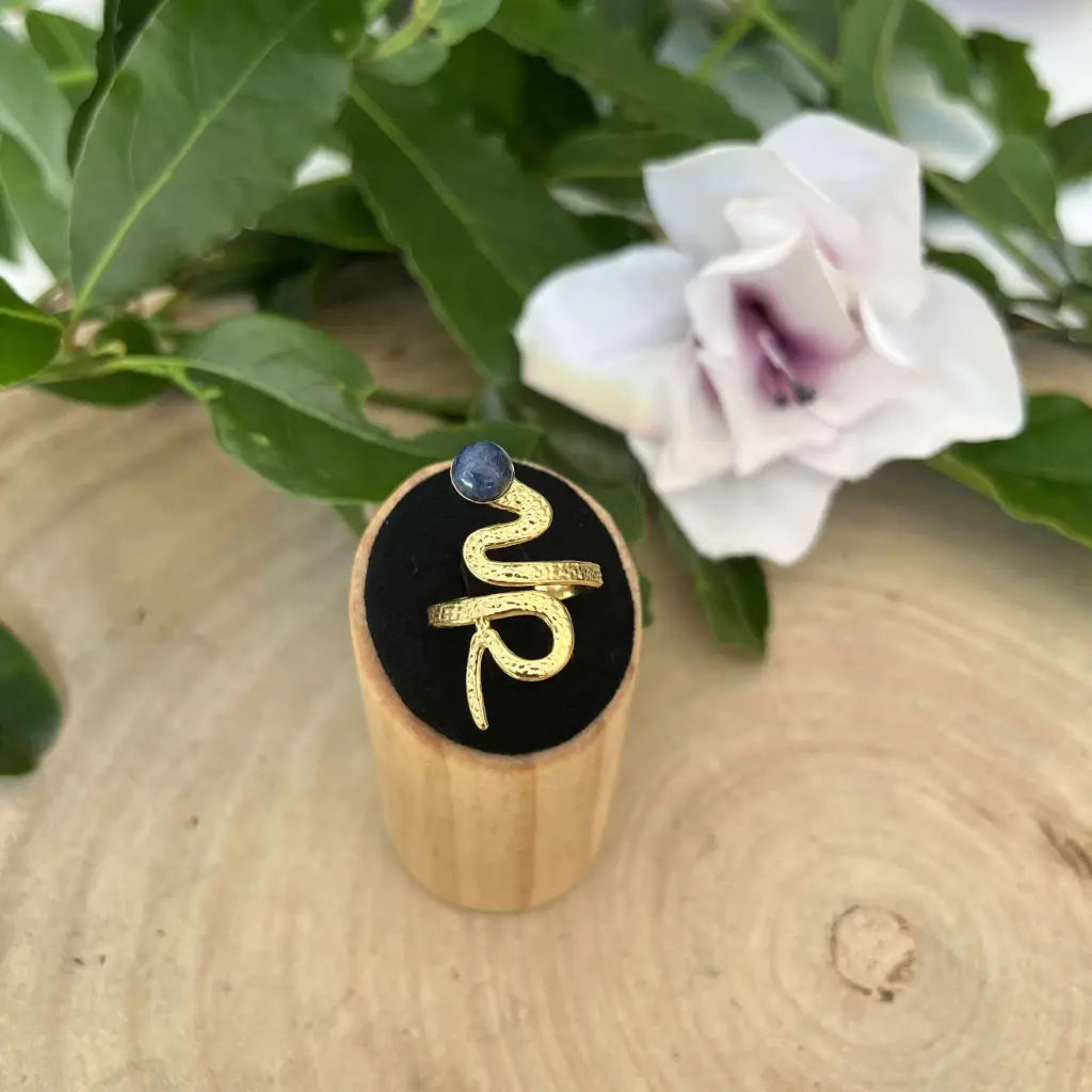 Bague ajustable Serpent dorée avec pierre bleue présentée sur support en bois près d'une fleur blanche