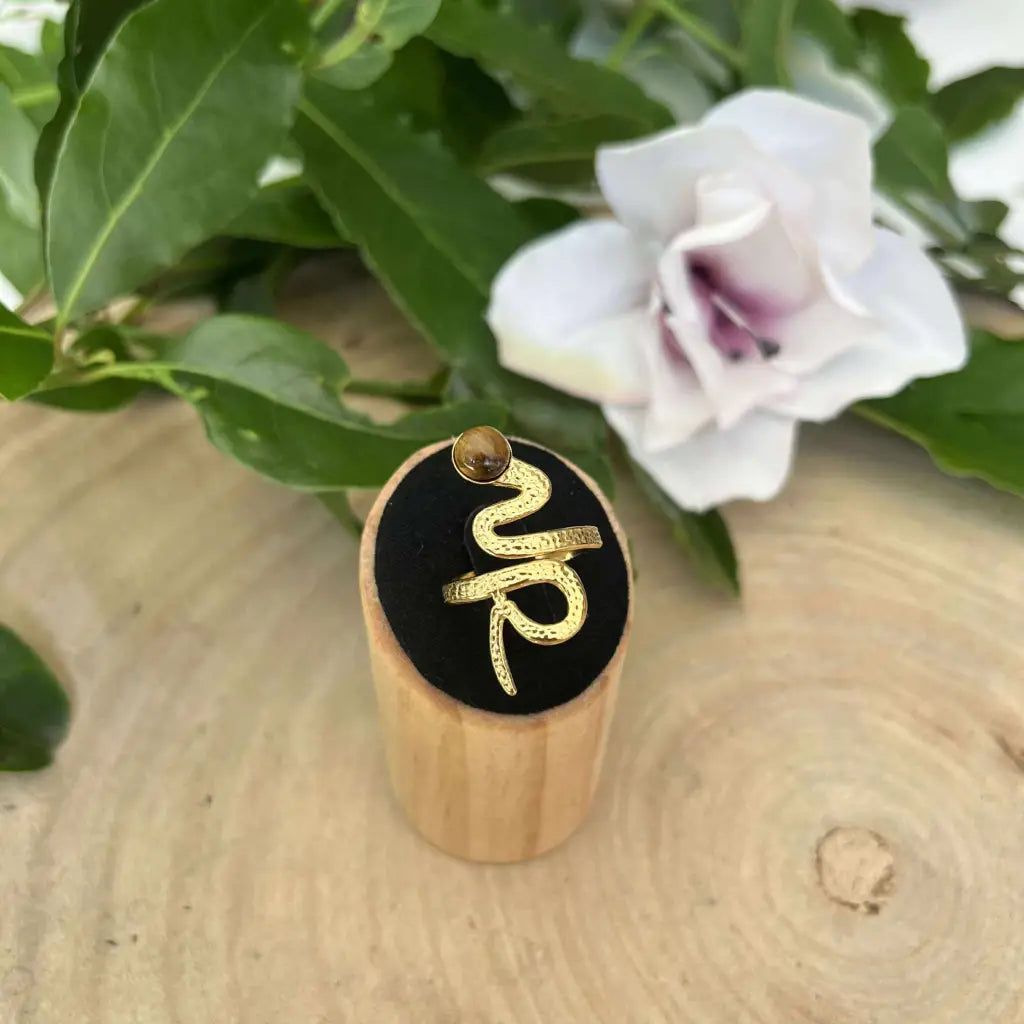 Bague ajustable Serpent dorée avec pierre brun clair présentée sur support en bois face à une fleur blanche