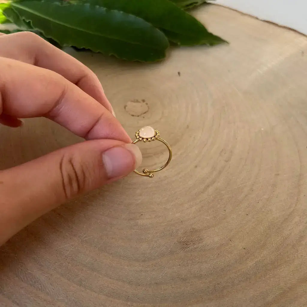Bague ajustable Solara en or tenue entre les doigts sur un fond en bois avec feuilles vertes