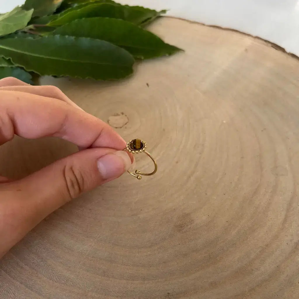 Bague ajustable Solara en or avec pierre sombre tenue à la main sur fond bois et feuilles vertes