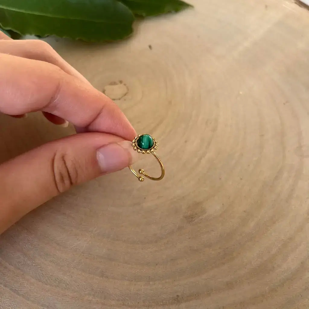 Bague ajustable Solara en or avec pierre verte tenue entre deux doigts sur fond en bois