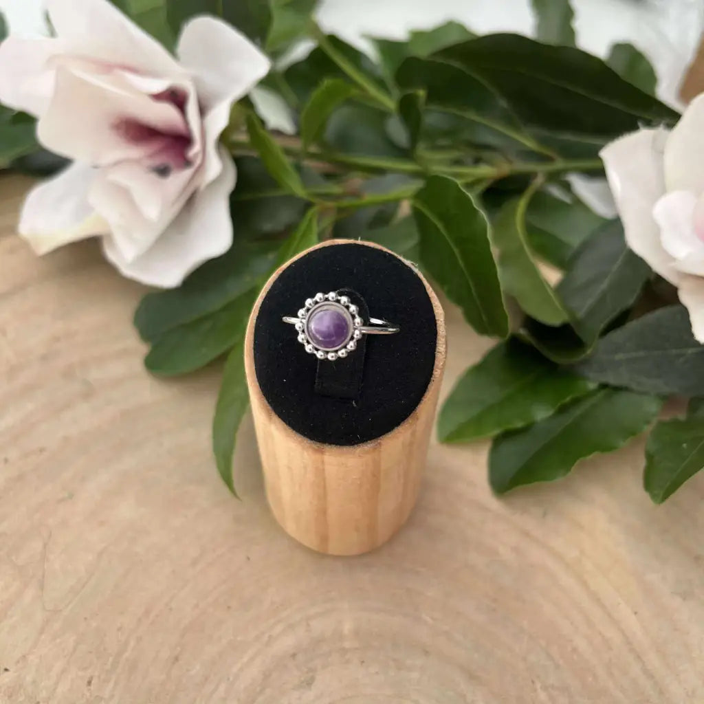 Bague ajustable Solara avec pierre violette sertie d'argent sur support en bois et fleurs en arrière-plan