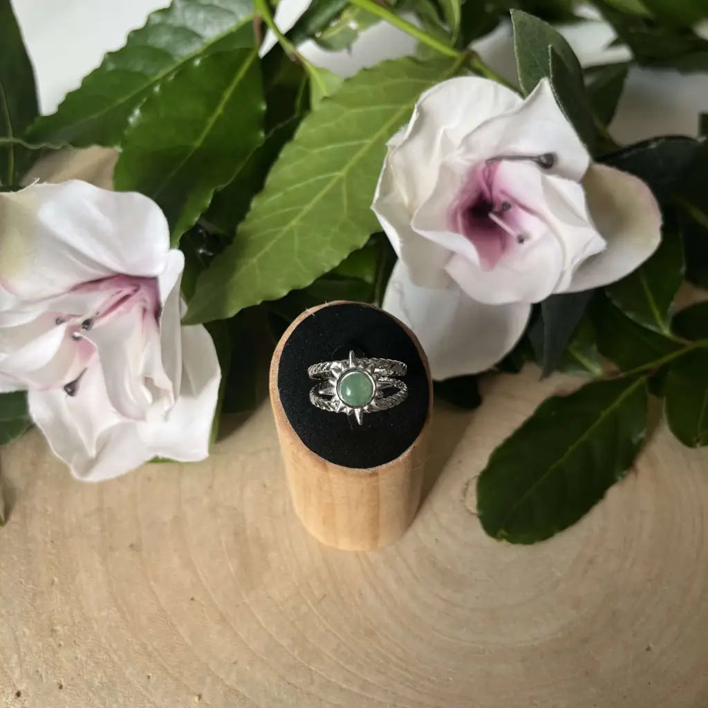 Bague ajustable Soleil en argent avec pierre verte présentée sur socle en bois entourée de fleurs blanches
