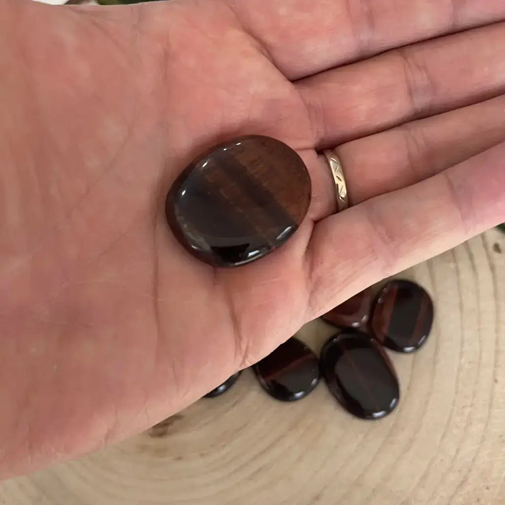 Galet Œil de Taureau poli brun rouge posé dans une main, autres galets brillants sur planche en bois Aura de Roche