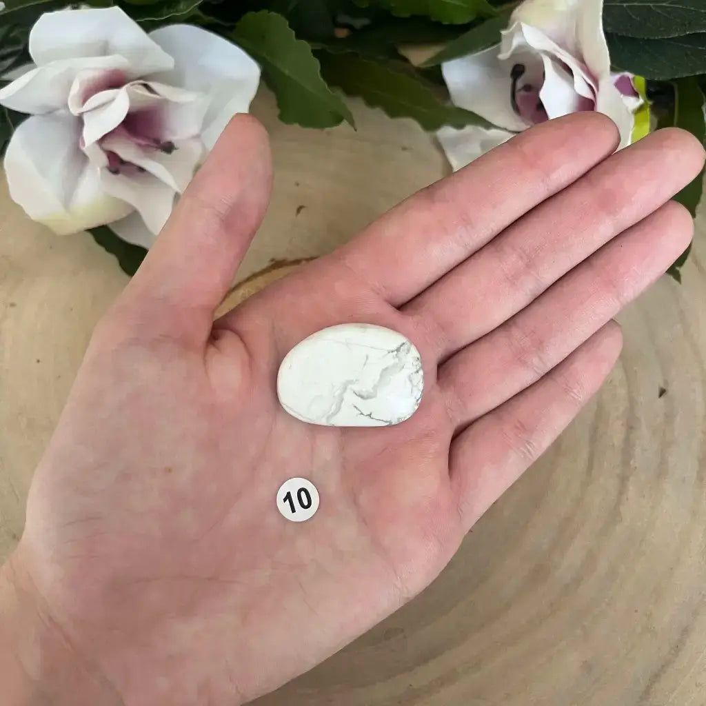 Galet Plat Howlite Blanche dans une main sur fond bois avec etiquette 10 et fleurs decoratives