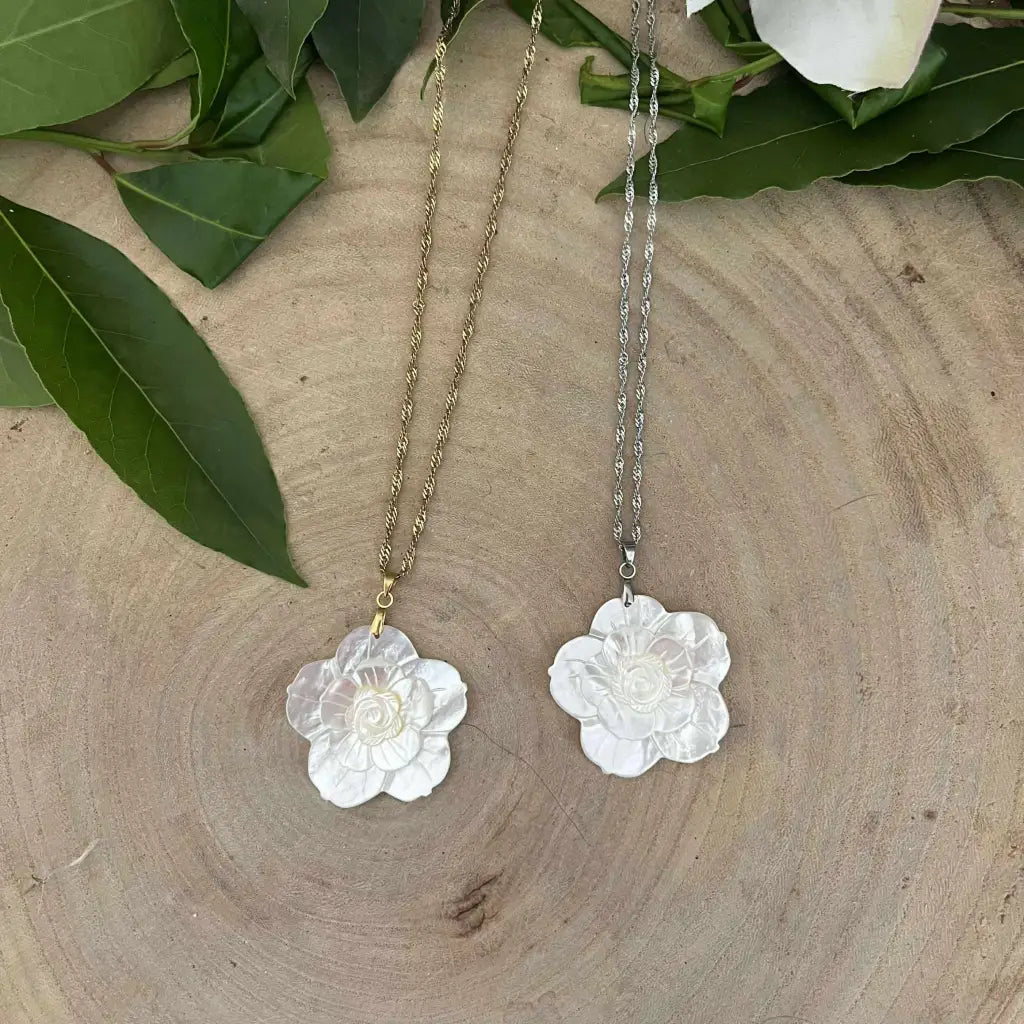 Pendentif Fleur en Nacre sur chaînes en or et argent posé sur fond bois naturel avec feuilles vertes