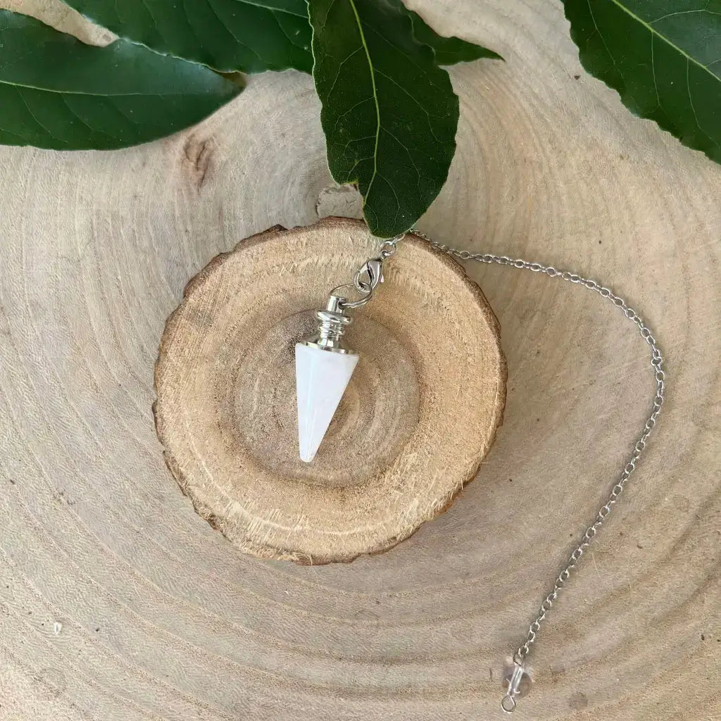 Pendule en Cristal de Roche blanc en forme de cône sur chaîne argentée présenté sur tranche de bois avec feuilles