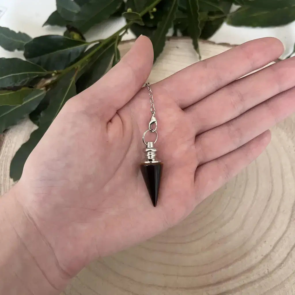 Pendule en Oeil de Tigre conique à chaîne et monture argent tenue dans la paume sur fond bois et feuilles