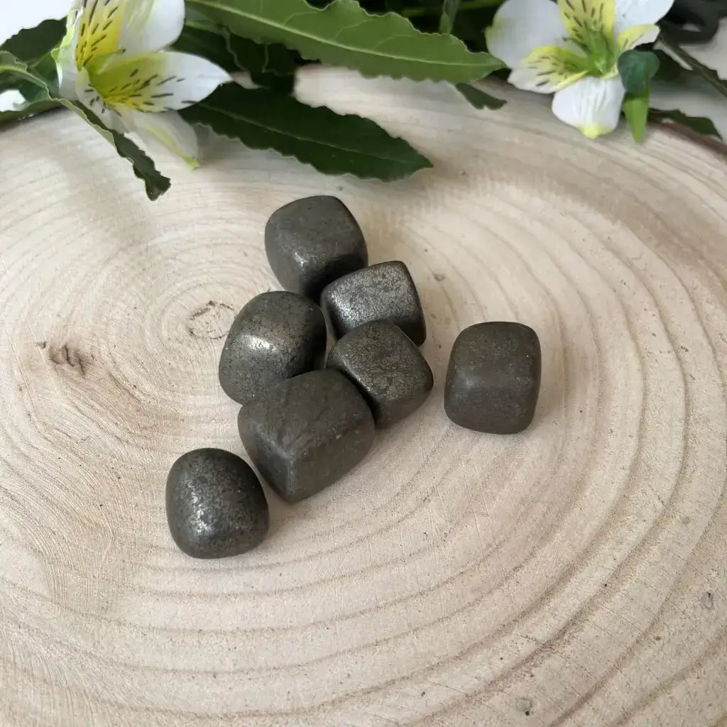 Sept pierres roulées Pyrite Naturelle posées sur un fond en bois avec des fleurs blanches en arrière-plan